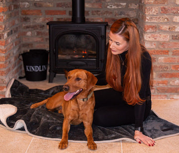 red fox labrador dog sat on grey 100% waterproof blanket, lady sat with dog, she has long ginger hair and wearing all black. They are sat infront of a log burner, brick on the walls and cream tiles. 