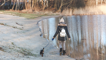 Lady walking her dog on the shore of beach, she wearing a white backpack, cream hat, green jacket. The dog is very small and theres trees in the background 